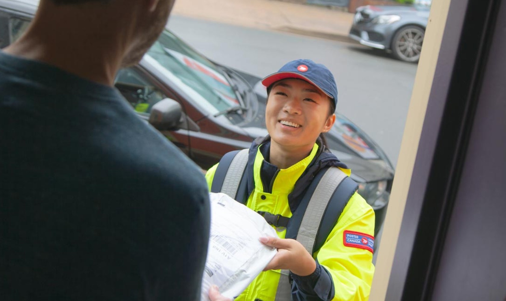 A Canada Post employee wearing a bright yellow jacket delivers a package to a man standing in his doorway.
