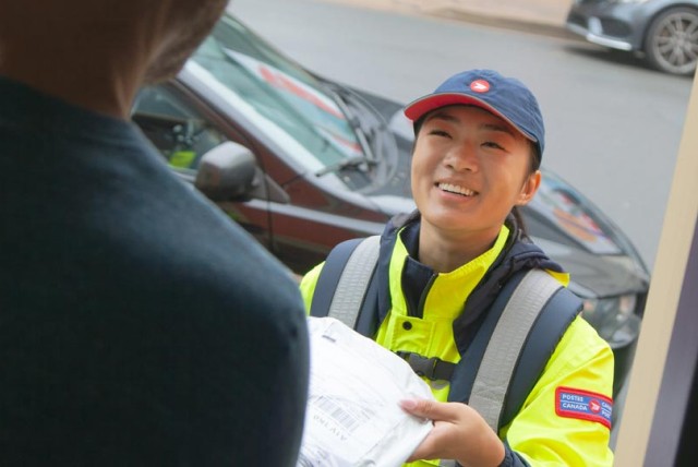 A Canada Post employee wearing a bright yellow jacket delivers a package to a man standing in his doorway.