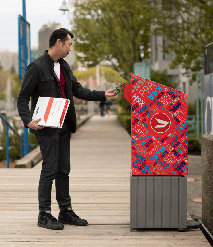 A man on a boardwalk opens the chute of a Canada Post Street letterbox. He holds a flat rate box in his other hand. A man on a boardwalk opens the chute of a Canada Post Street letterbox. He holds a flat rate box in his other hand.