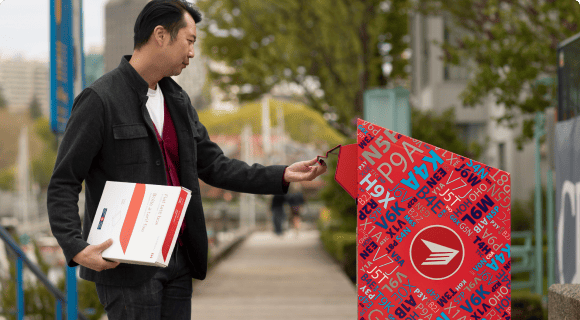 A man on a boardwalk opens the chute of a Canada Post Street letterbox. He holds a flat rate box in his other hand. A man on a boardwalk opens the chute of a Canada Post Street letterbox. He holds a flat rate box in his other hand.