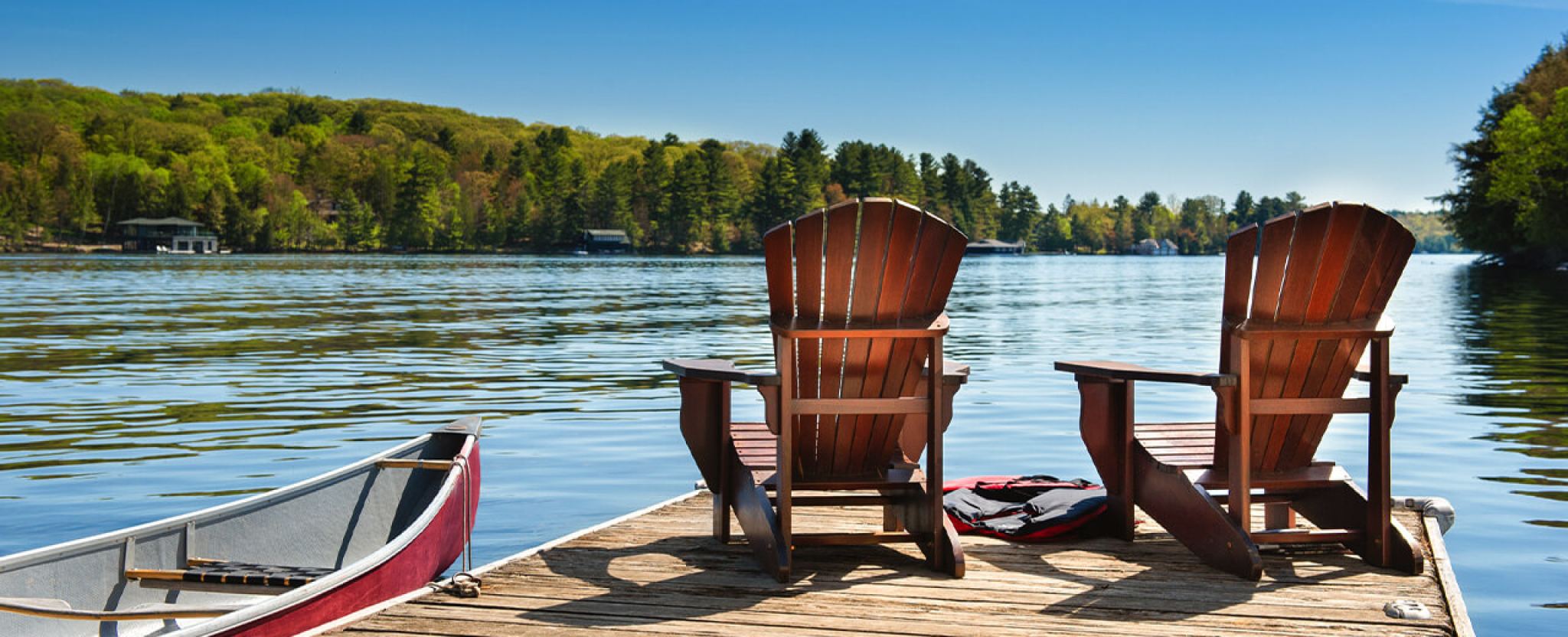 Two wooden chairs sit on a lakeside dock, next to a canoe.