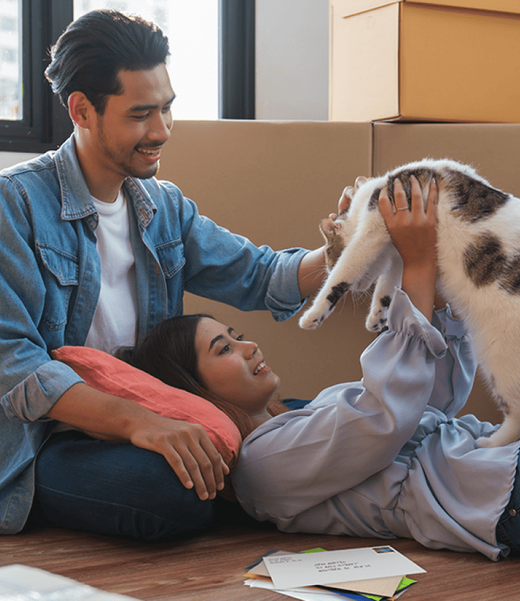 A young couple play with a cat on the floor of their home. Moving boxes are stacked behind them.