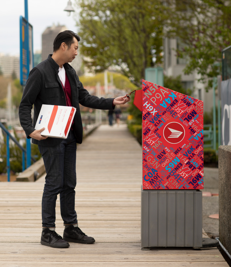 A man holding a flat rate box opens a Canada Post street mailbox. 