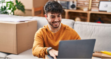 A person sits on a couch smiling at a laptop, surrounded by boxes, suggesting a recent move or online activity at home.