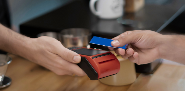 A hand holds a red payment terminal while another taps a blue card, showing a contactless transaction on a caf&eacute; counter.