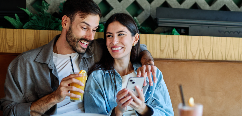 Two people sit close together in a caf&eacute;, one smiling at a smartphone, the other holding a drink, with plants in the background.