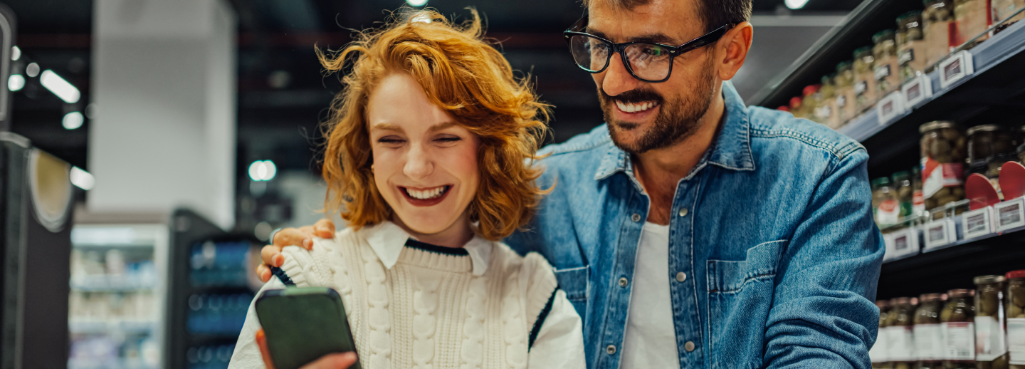 Two people stand behind a shopping cart in a grocery aisle, looking at a smartphone together with shelves in the background.