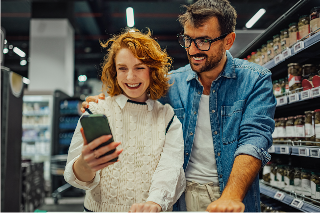 Two people stand behind a shopping cart in a grocery aisle, looking at a smartphone together with shelves in the background.