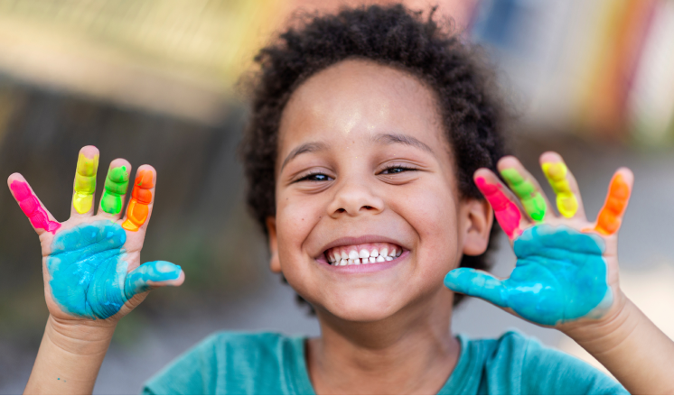 A young child grins while holding up their hands, each finger and palm painted a different colour. 