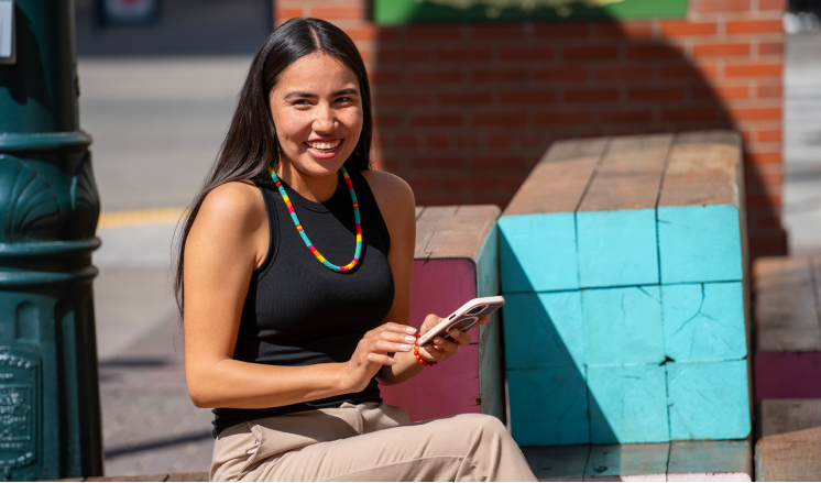 A woman sitting on a bench in an outdoor urban area, holding her smartphone and smiling.