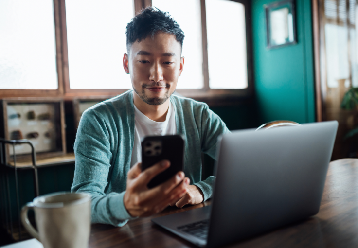 Un homme regarde son téléphone cellulaire alors qu’il est assis devant un ordinateur portable à une table en bois