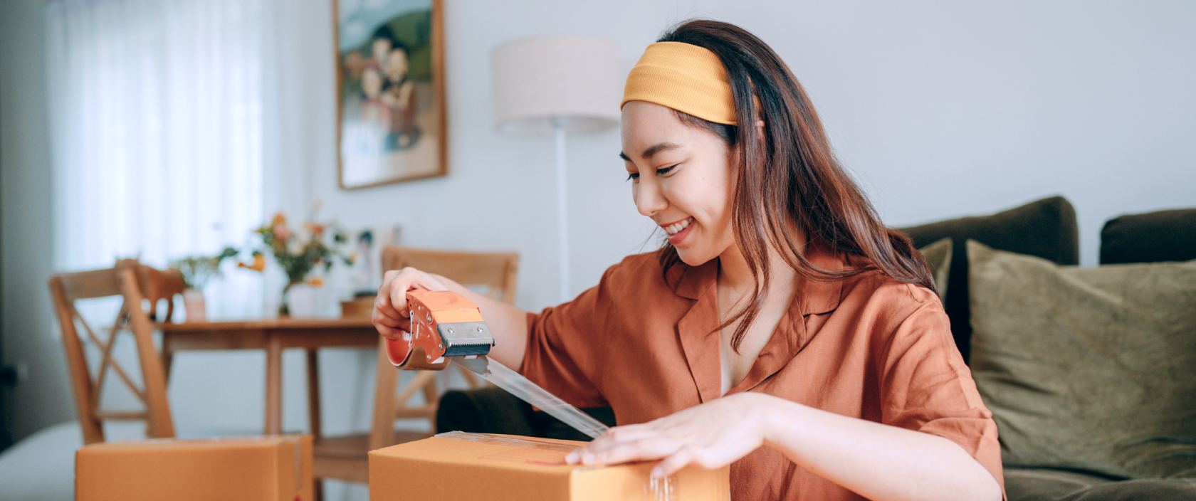 A young woman wearing a headband seals a shipping box with tape.