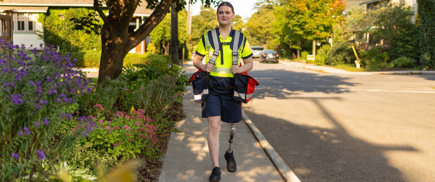 Une factrice de Postes Canada avec une jambe prothétique marche dans une rue résidentielle en portant des sacs de courrier. 