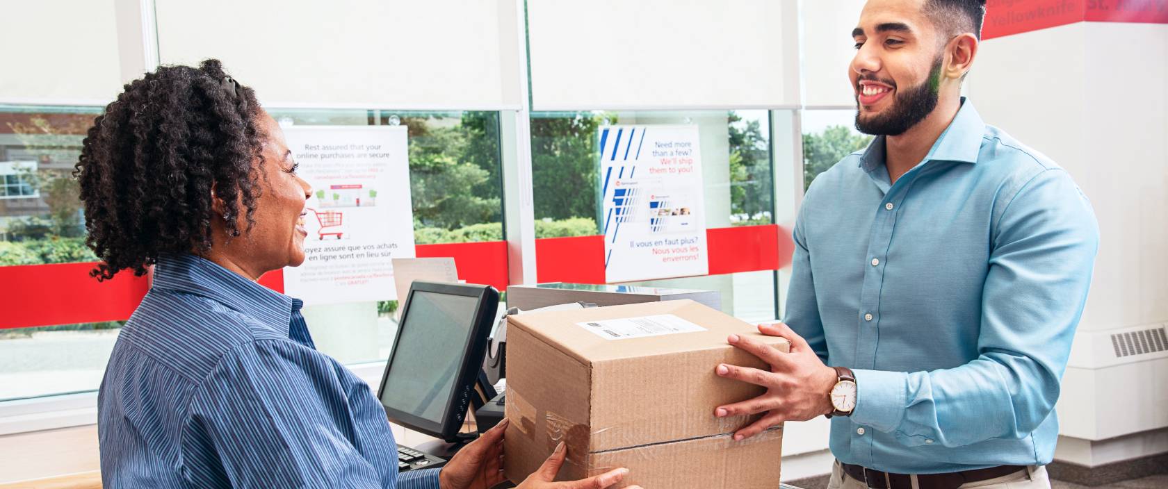 Une employée souriante d’un bureau de poste de Postes Canada remet un colis à un homme qui se trouve de l’autre côté du comptoir