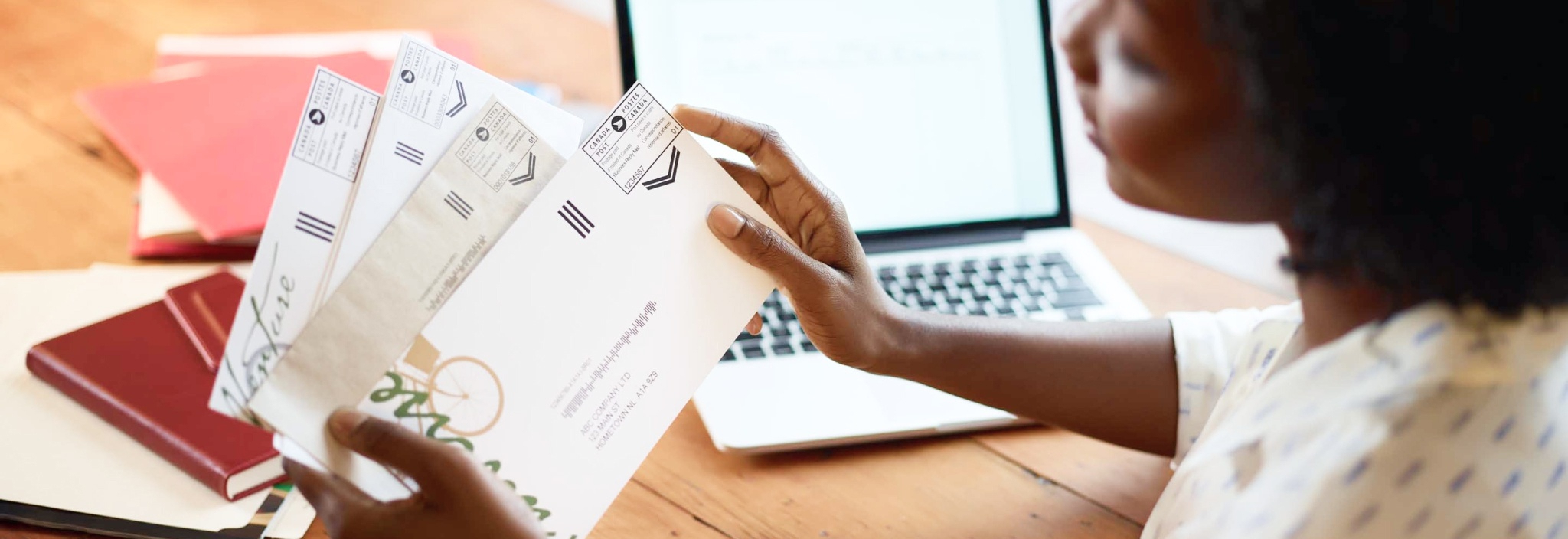 Une femme à son bureau tient du courrier d’affaires. Il y a une vignette postale de Postes Canada dans le coin supérieur droit de chaque enveloppe.