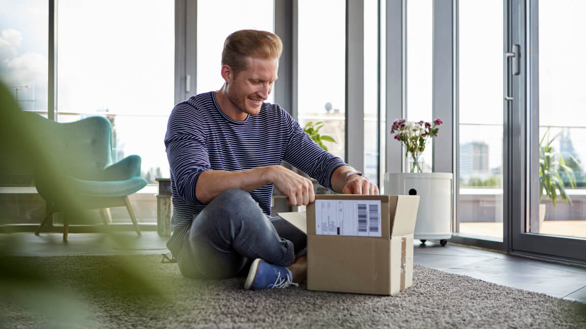 Un homme est assis sur le plancher d’un appartement devant une boîte ouverte comportant une étiquette d’expédition.