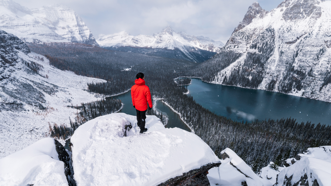 Vue aérienne d’une personne en manteau rouge sur une montagne enneigée surplombant une vallée boisée avec un lac bleu.