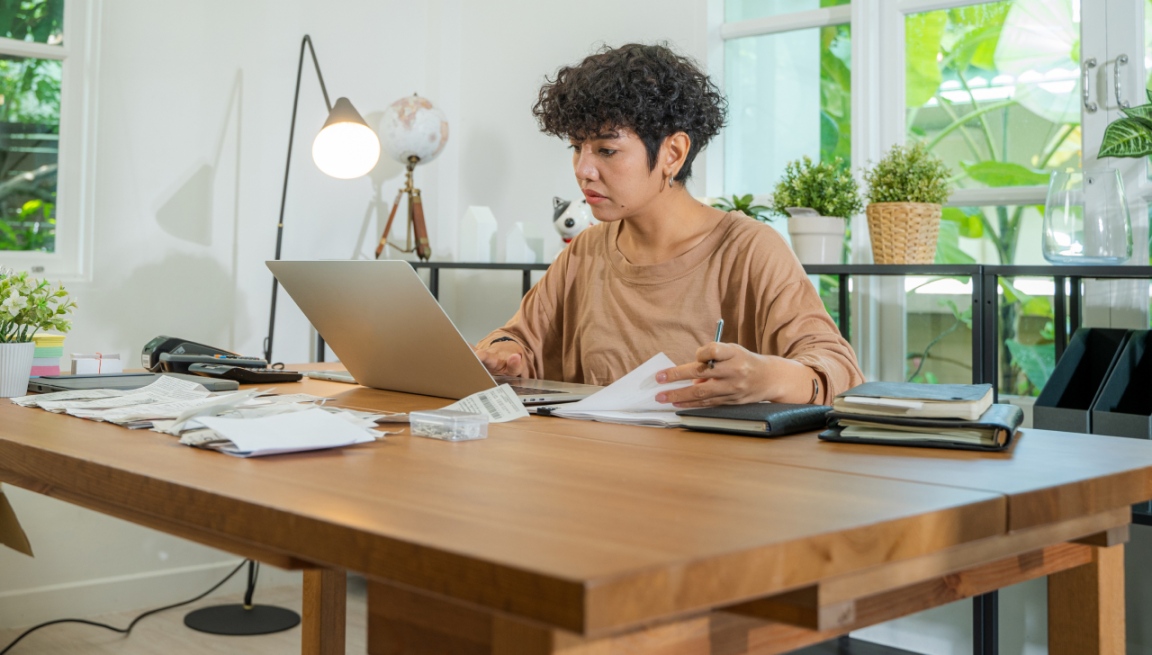 Une femme est assise à un bureau où elle travaille sur un ordinateur portable en feuilletant des documents.