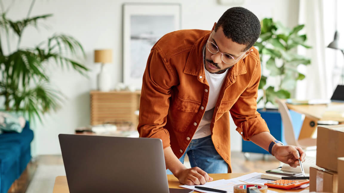 Un homme penché au-dessus d’un bureau couvert de papier et d’articles d’emballage regarde l’écran d’un ordinateur portable. 