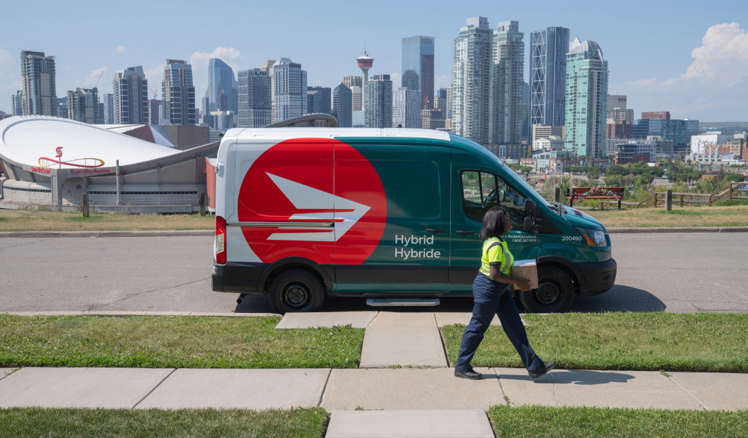 Une camionnette hybride de Postes Canada stationnée sur la rue avec en arrière plan, la silhouette du centre-ville de Calgary. Sur le trottoir, une employée de Postes Canada transporte un colis dans ses mains.